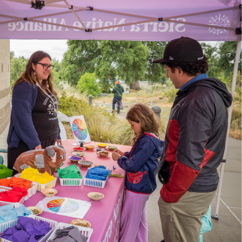 An adult man and young girl interact with a colorful vendor booth and the woman behind it. 